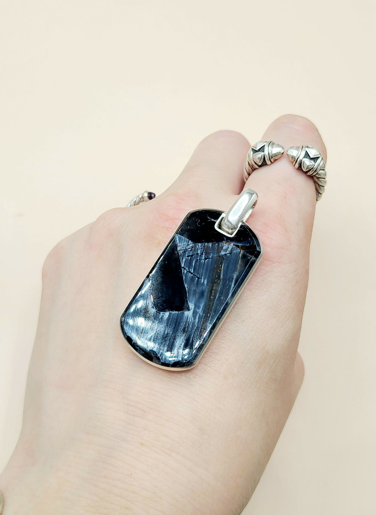A close-up of a hand wearing a large rectangular dark blue stone pendant with a silver-toned metal frame and bail, alongside textured silver rings on the fingers, against a plain light beige background.