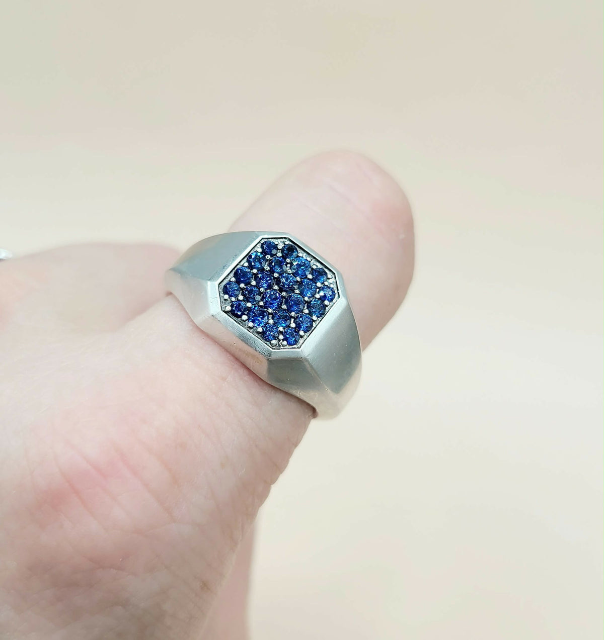 A silver-toned signet ring with a faceted octagonal face paved with deep blue stones, shown worn on a finger against a soft beige background.