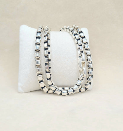 Three silver-toned box-link chains with clear rectangular gemstones, displayed on a white jewelry stand against a light beige background.