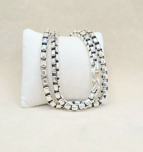 Three silver-toned box-link chains with clear rectangular gemstones, displayed on a white jewelry stand against a light beige background.