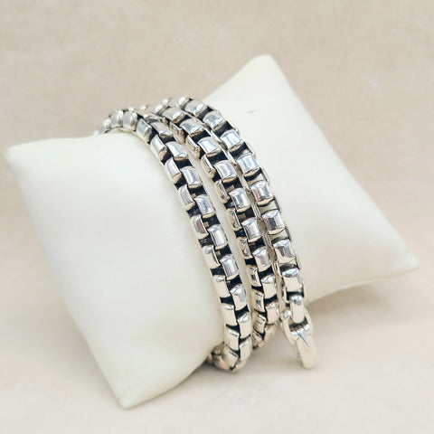 Three silver-toned chain link bracelets stacked on a white display pillow against a light beige background.
