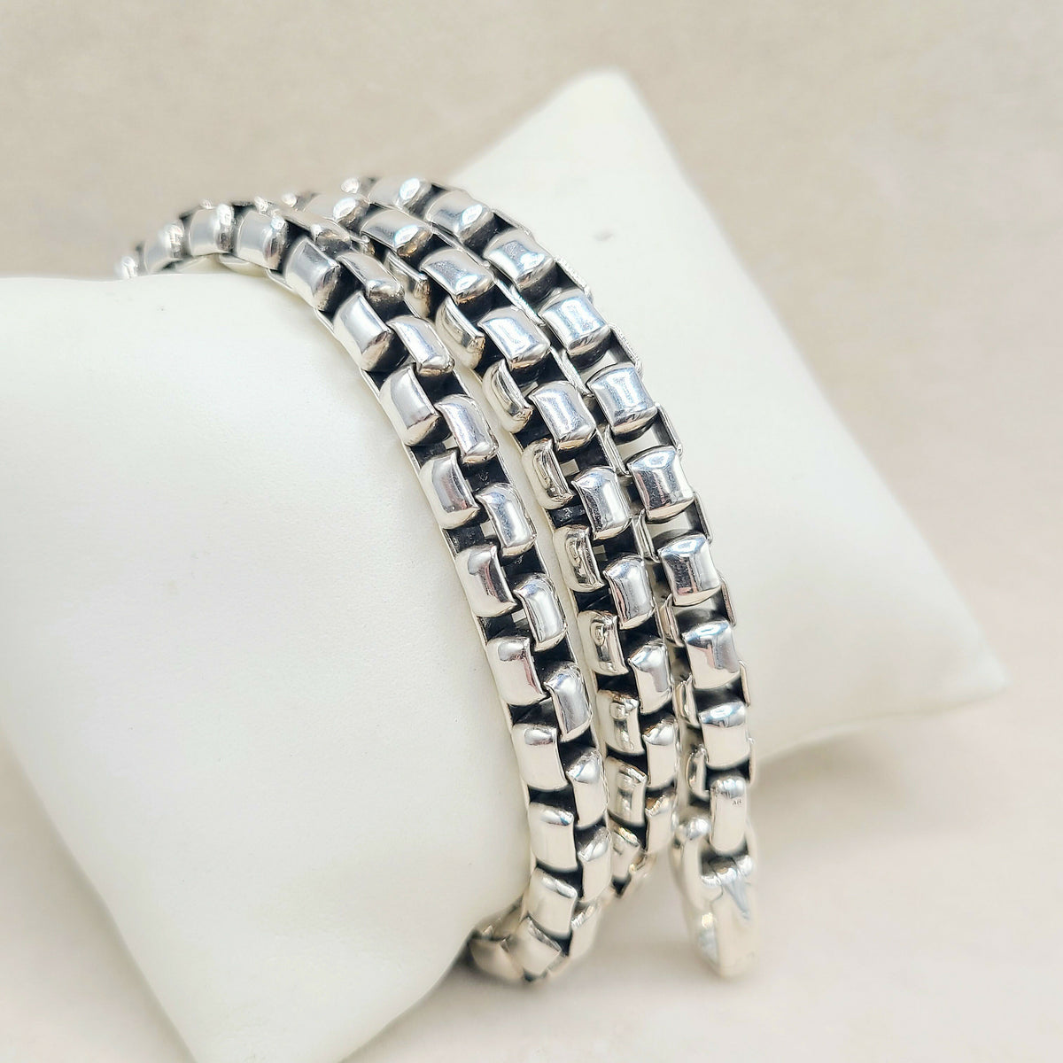 Three silver-toned box-link chain bracelets stacked on a white display stand against a light beige background.