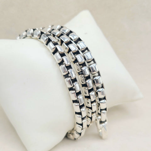Three silver-toned box-link chain bracelets stacked on a white display stand against a light beige background.