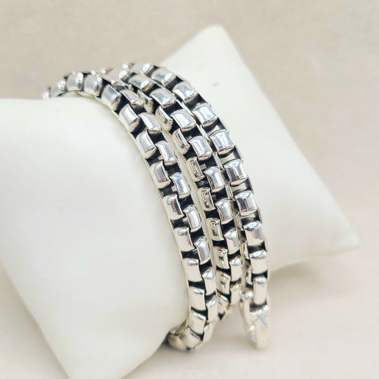 Three silver-toned box-link chain bracelets stacked on a white display stand against a light beige background.