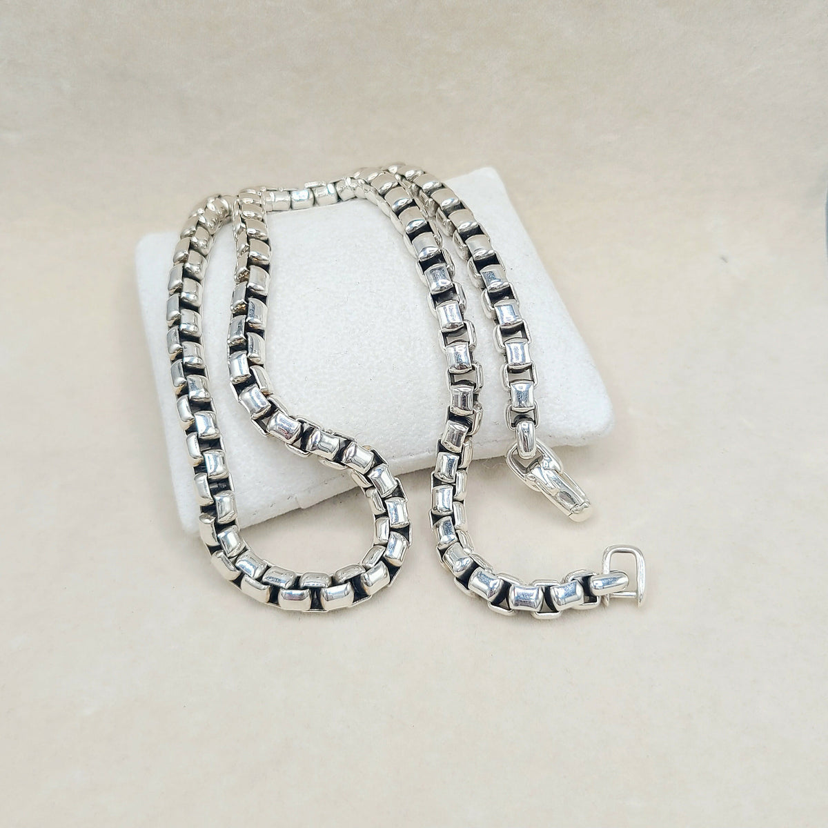 A silver-toned box chain necklace with clear rectangular crystals, draped over a white display cushion on a light beige background.