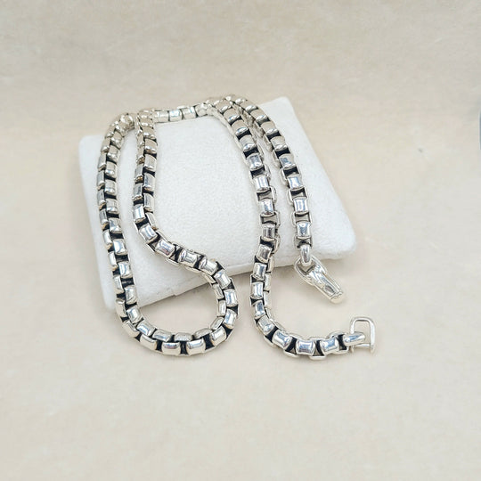 A silver-toned box chain necklace with clear rectangular crystals, draped over a white display cushion on a light beige background.
