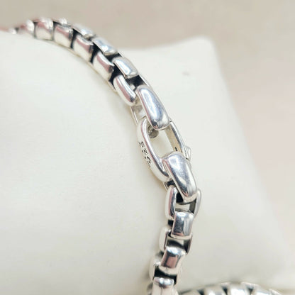 A close-up of a polished silver-toned metal chain bracelet displayed on a white jewelry stand, showing the reflective links and a small stamped '925' mark.