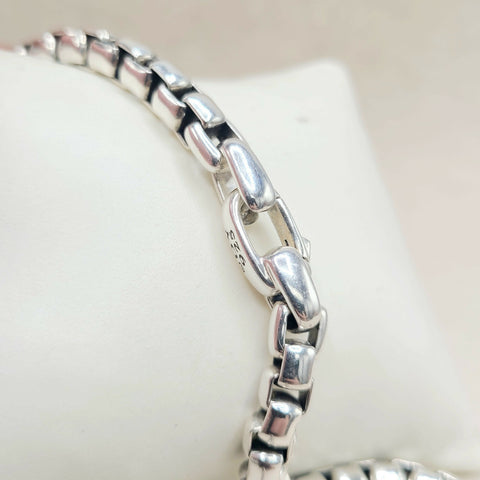 A close-up of a polished silver-toned metal chain bracelet displayed on a white jewelry stand, showing the reflective links and a small stamped '925' mark.