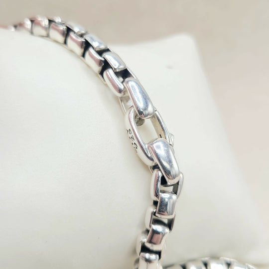 A close-up of a polished silver-toned metal chain bracelet displayed on a white jewelry stand, showing the reflective links and a small stamped '925' mark.