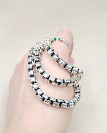 A close-up of a hand wearing a chunky silver chain bracelet with clear rectangular stones, alongside a thin silver ring with small green stones, against a light beige background.