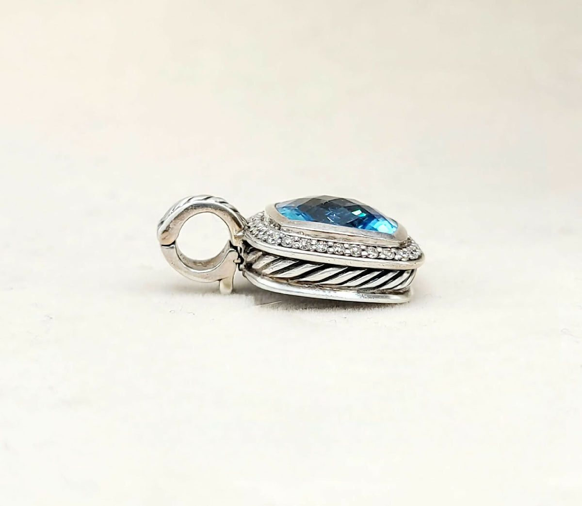 A sterling silver pendant charm featuring a teardrop-shaped blue topaz stone surrounded by small clear pavé stones, with a textured twisted border and a hanging loop, set against a plain off-white background.