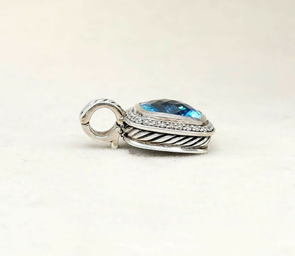 A sterling silver pendant charm featuring a teardrop-shaped blue topaz stone surrounded by small clear pavé stones, with a textured twisted border and a hanging loop, set against a plain off-white background.