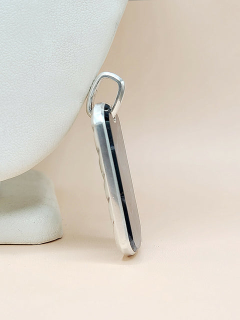 A polished silver rectangular pendant with a rounded top loop, shown standing vertically on a light beige surface next to a white textured display stand.