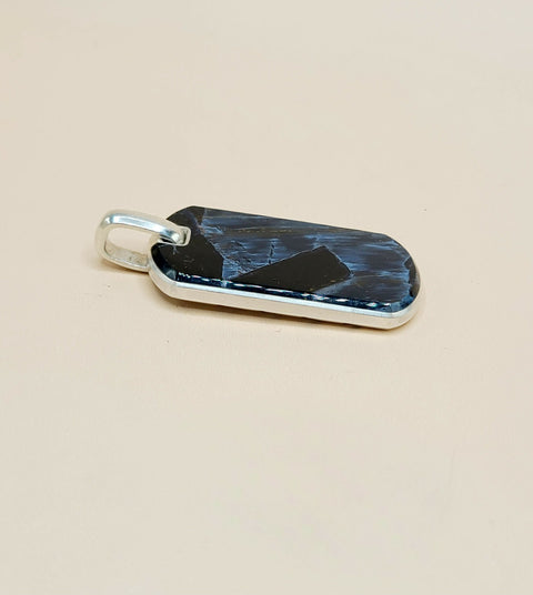 A rectangular pendant with rounded corners, featuring a polished dark blue and black marbled or textured face, set in a silver-tone metal bezel with a silver bail, shown on a plain beige background.