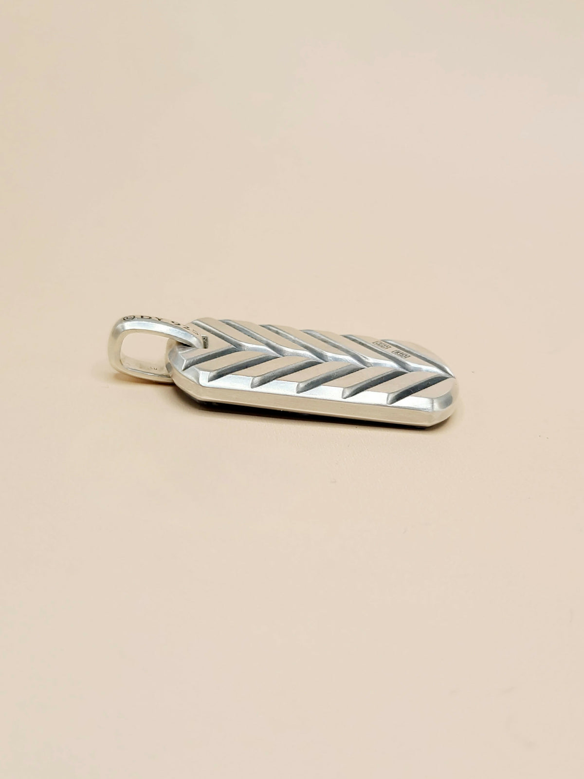 A silver chevron-patterned pendant with a small loop at the top, shown on a plain beige background.
