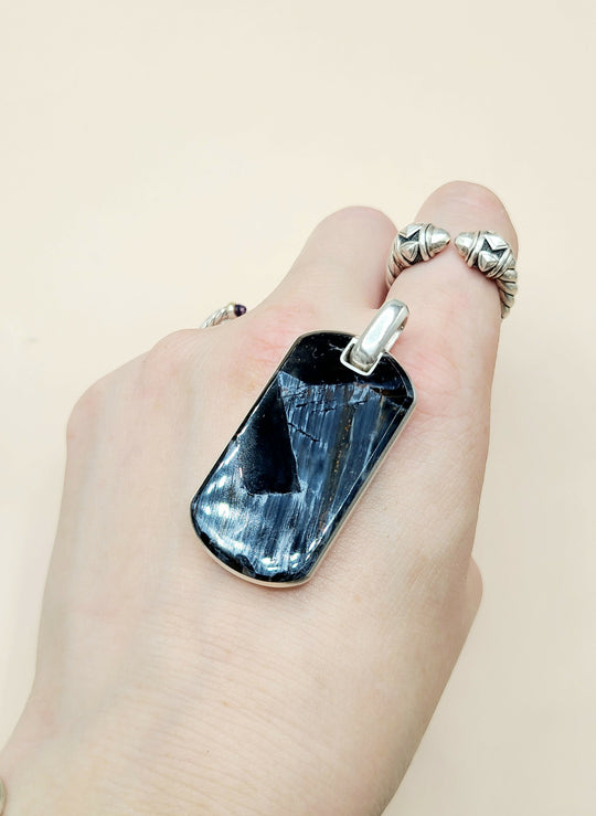 A close-up of a hand wearing a large rectangular dark blue stone pendant with a silver-toned metal frame and bail, alongside textured silver rings on the fingers, against a plain light beige background.
