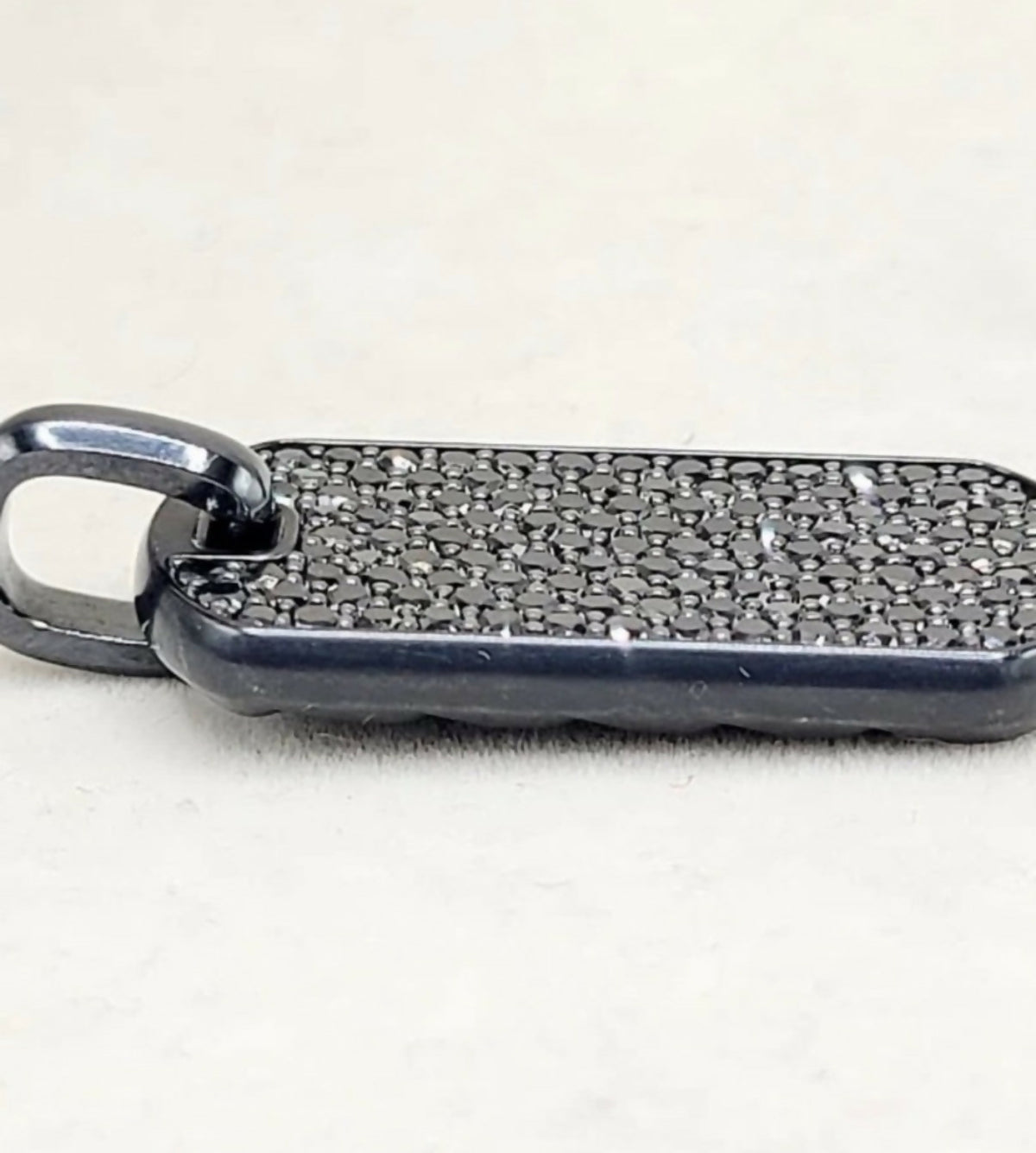 A close-up of a black metal pendant or tag with a textured, pebble-like surface and a small attached loop, photographed on a light off-white background.