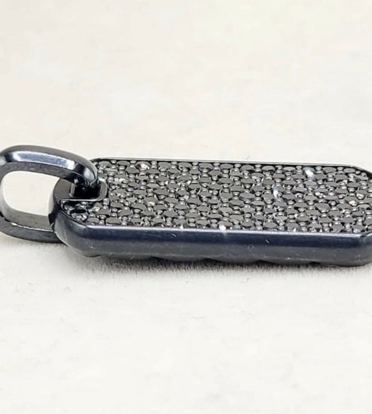 A close-up of a black metal pendant or tag with a textured, pebble-like surface and a small attached loop, photographed on a light off-white background.