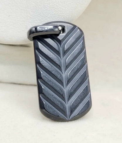 A dark gray textured leather dog tag pendant with a leaf pattern and a silver-tone metal loop at the top, resting on a light beige surface.