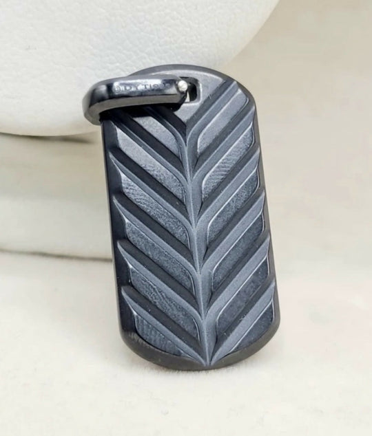 A dark gray textured leather dog tag pendant with a leaf pattern and a silver-tone metal loop at the top, resting on a light beige surface.