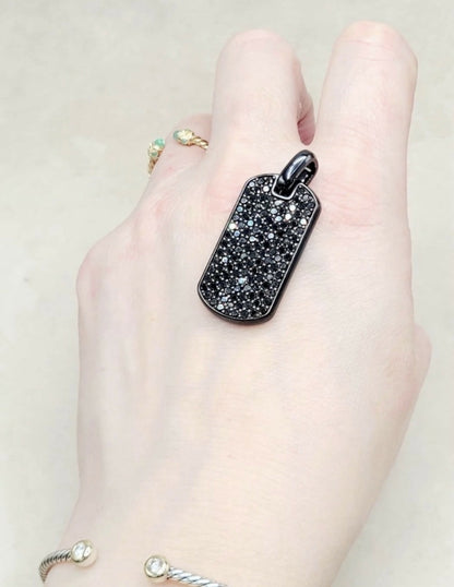 A close-up of a hand wearing a black dog tag pendant covered in small clear stones, with other rings and bracelets also visible on the hand against a light beige background.