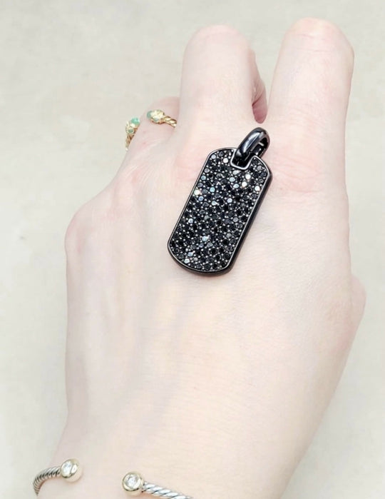 A close-up of a hand wearing a black dog tag pendant covered in small clear stones, with other rings and bracelets also visible on the hand against a light beige background.