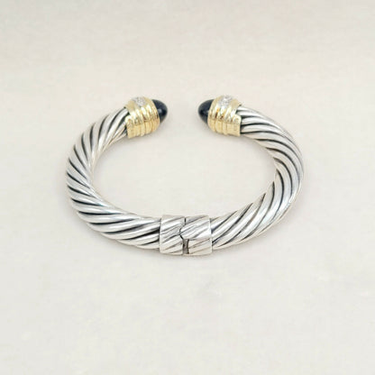A silver cable-style cuff bracelet with twisted metallic bands, gold-toned end caps, and small dark stone accents at the tips, photographed on a plain light background.