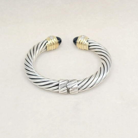A silver cable-style cuff bracelet with twisted metallic bands, gold-toned end caps, and small dark stone accents at the tips, photographed on a plain light background.