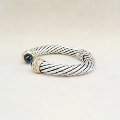 A silver cable-style cuff bracelet with gold-toned end caps, one featuring a dark blue cabochon stone and the other accented with small clear stones, photographed on a light beige background.