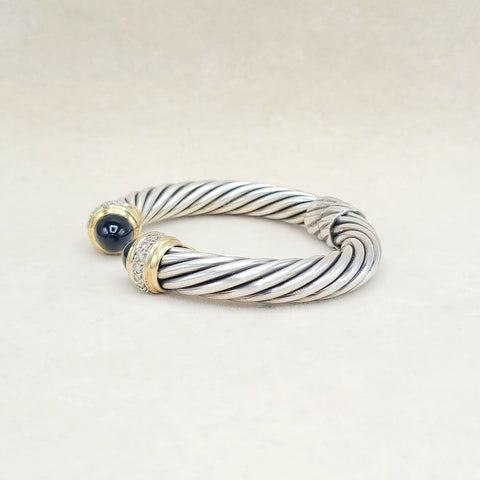 A silver cable-style cuff bracelet with gold-toned end caps, one featuring a dark blue cabochon stone and the other accented with small clear stones, photographed on a light beige background.