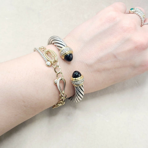 A close-up of a person's wrist wearing three bracelets: a silver-toned twisted cable bracelet with a black onyx bead, a gold-toned chain bracelet with a gold-toned lobster clasp, and a silver-toned twisted cable bracelet with a black onyx bead and crystal detailing.