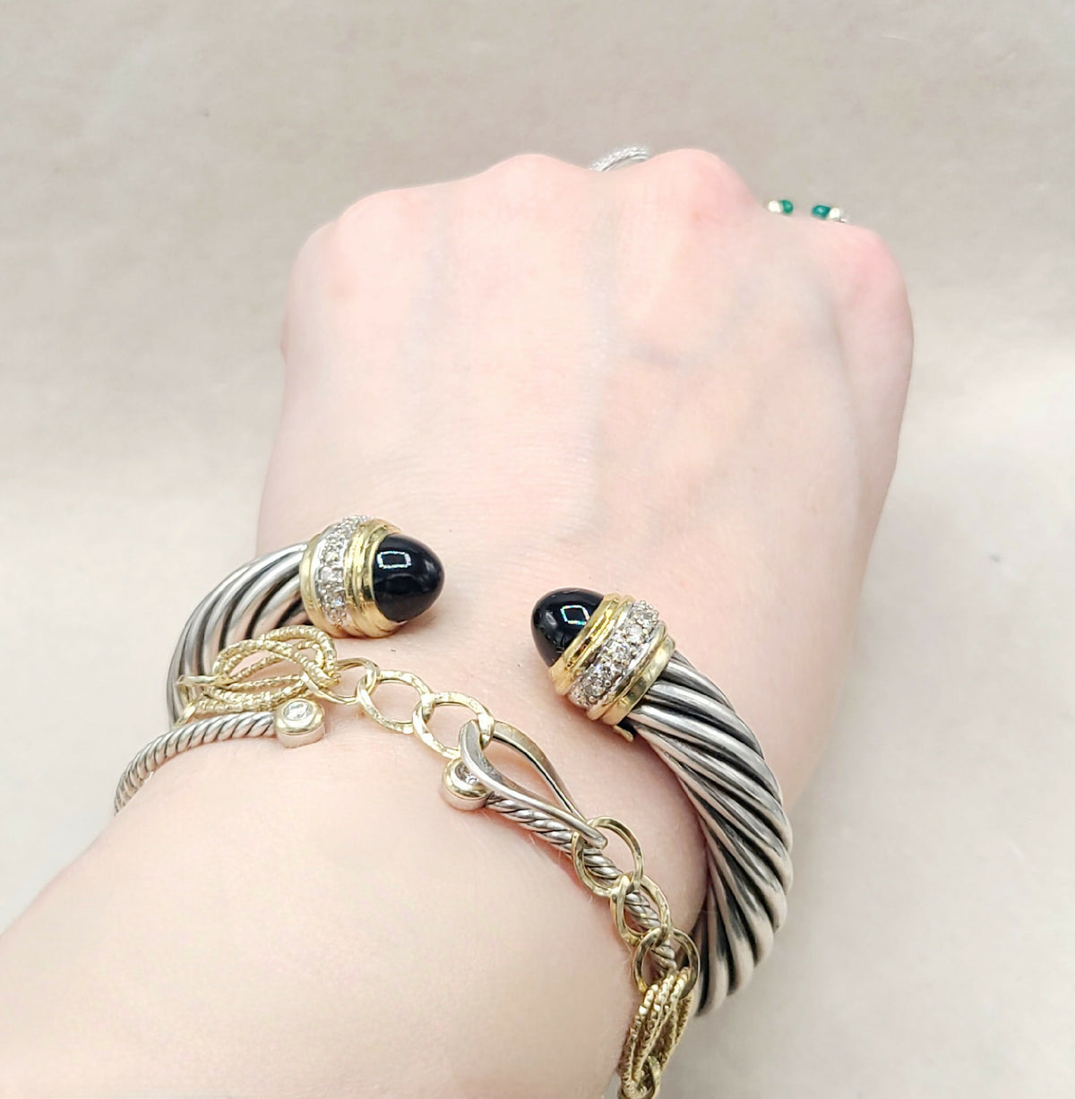 A close-up of a person's wrist wearing multiple bracelets, including a silver and black twisted cable bracelet with black cabochon stones, a gold and silver rope bracelet, and a gold chain bracelet with a small clear stone.