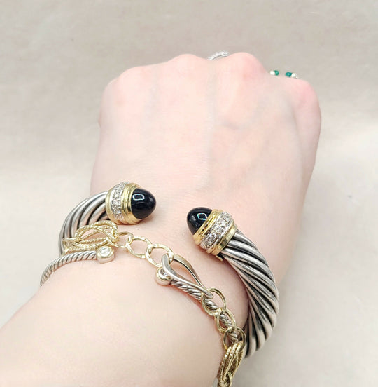A close-up of a person's wrist wearing multiple bracelets, including a silver and black twisted cable bracelet with black cabochon stones, a gold and silver rope bracelet, and a gold chain bracelet with a small clear stone.