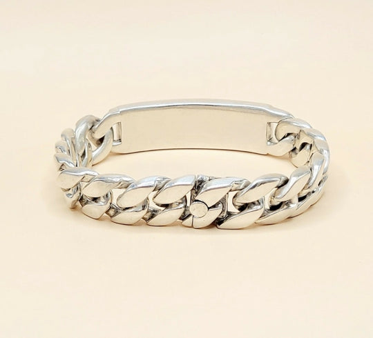 A silver-toned metal ID bracelet with a polished curb link design and a smooth rectangular ID plate, shown on a light beige background.