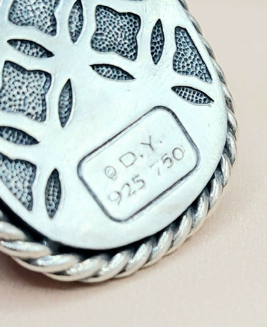 Close-up of a silver pendant with intricate design and visible hallmark on a beige background