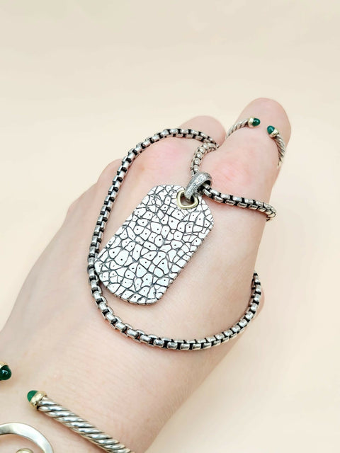 A close-up of a hand wearing a silver ring with a dog tag pendant featuring a crackle oxidized finish, paired with a matching silver beaded chain and small green stone accents.