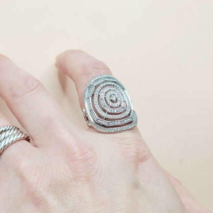 A silver spiral ring with pavé-set clear stones worn on a finger, with a second silver band visible on another finger, against a plain light beige background.