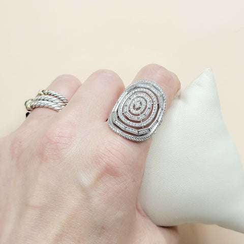 A silver-toned ring with a large square face and concentric spiral pattern set with small clear stones, shown worn on a hand next to a white display cushion.