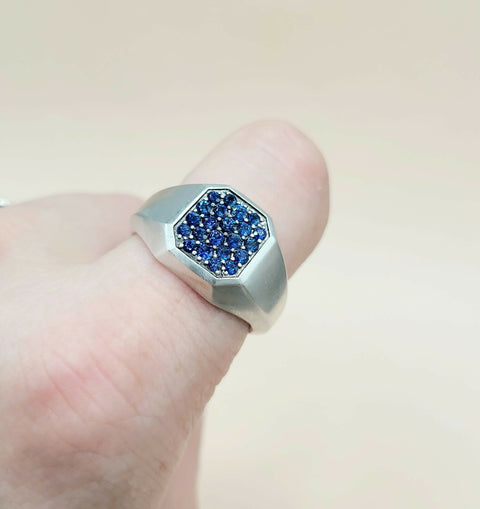 A silver-toned signet ring with a faceted octagonal face paved with deep blue stones, shown worn on a finger against a soft beige background.