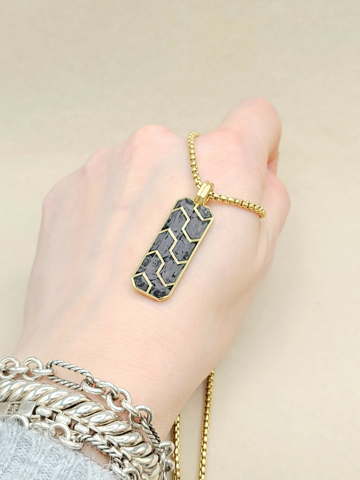 Hand wearing a gold chain with a hexagonal pendant on a beige background