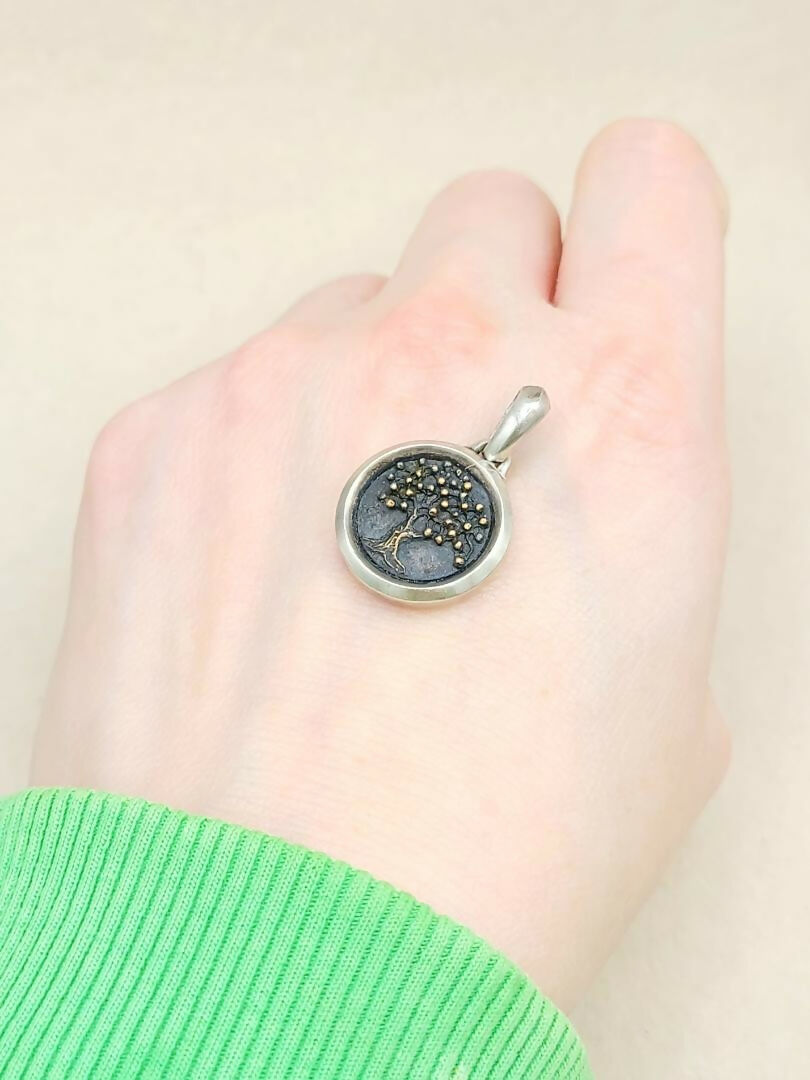 Silver pendant with tree design held in a hand against a beige background