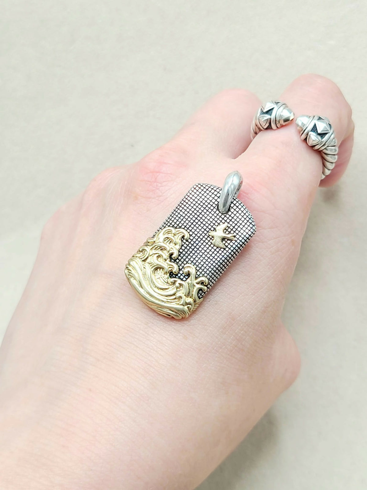 A close-up of a hand wearing a silver and gold ring. The ring features a rectangular textured silver plate with a gold wave design along the bottom edge and a small gold cross, with a silver bail at the top.