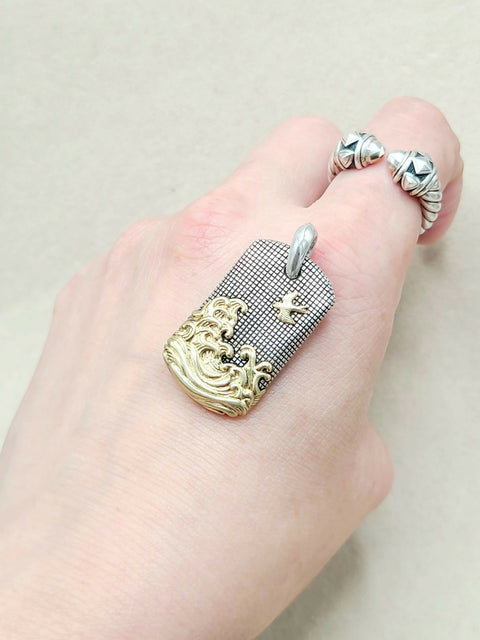 A close-up of a hand wearing a silver and gold ring. The ring features a rectangular textured silver plate with a gold wave design along the bottom edge and a small gold cross, with a silver bail at the top.