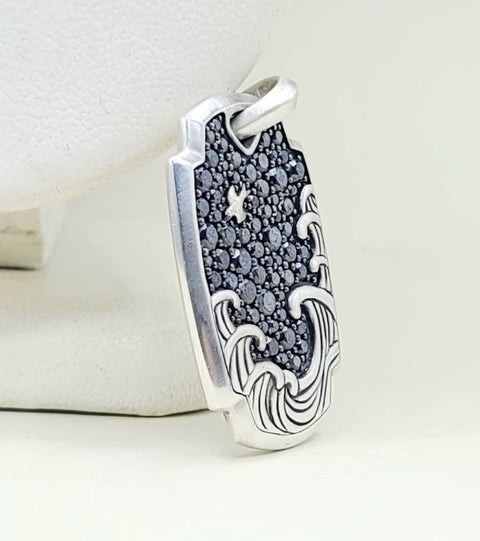 A sterling silver pendant featuring a wave design with a pavé black stone center and a small star motif, displayed on a white jewelry stand.