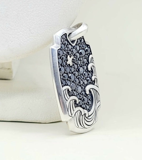 A sterling silver pendant featuring a wave design with a pavé black stone center and a small star motif, displayed on a white jewelry stand.