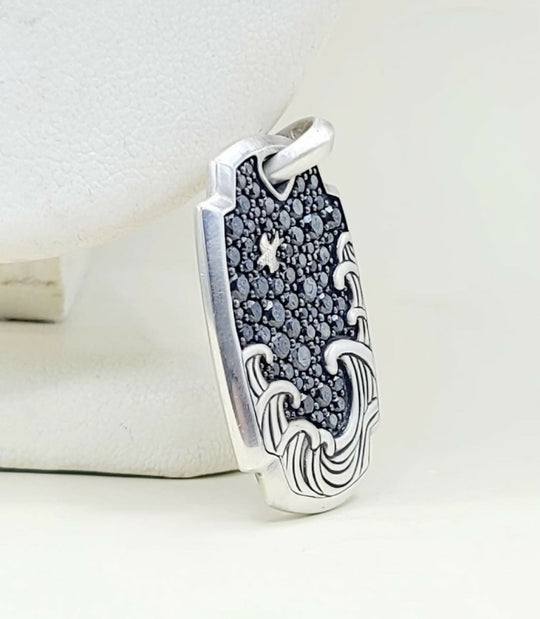 A silver pendant featuring a wave design with a dark, textured center and a small star motif, displayed on a white jewelry stand.