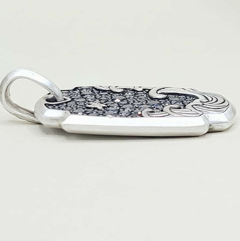 A sterling silver pendant with a polished bail and a textured, patterned top surface featuring dark inlay and engraved details, shown on a neutral light gray background.