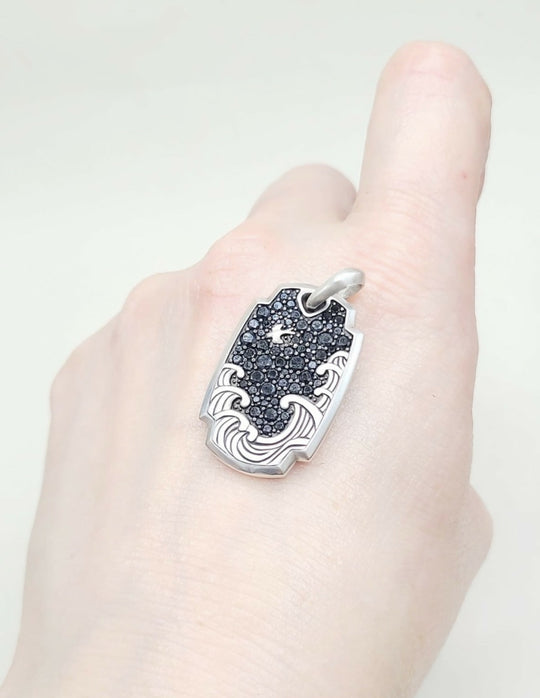 A silver-toned pendant with a black textured center and wave-like cutouts, shown worn on a person's finger against a light background.