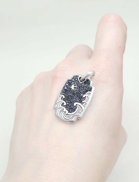 A sterling silver pendant with a black starry sky and wave design, shown worn on a finger.