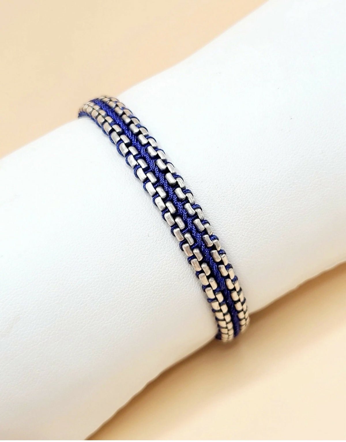 A close-up of a double-strand bracelet with a silver-toned curb link chain and a royal blue cord wrapped around it, displayed on a white cylindrical mannequin wrist against a soft beige background.